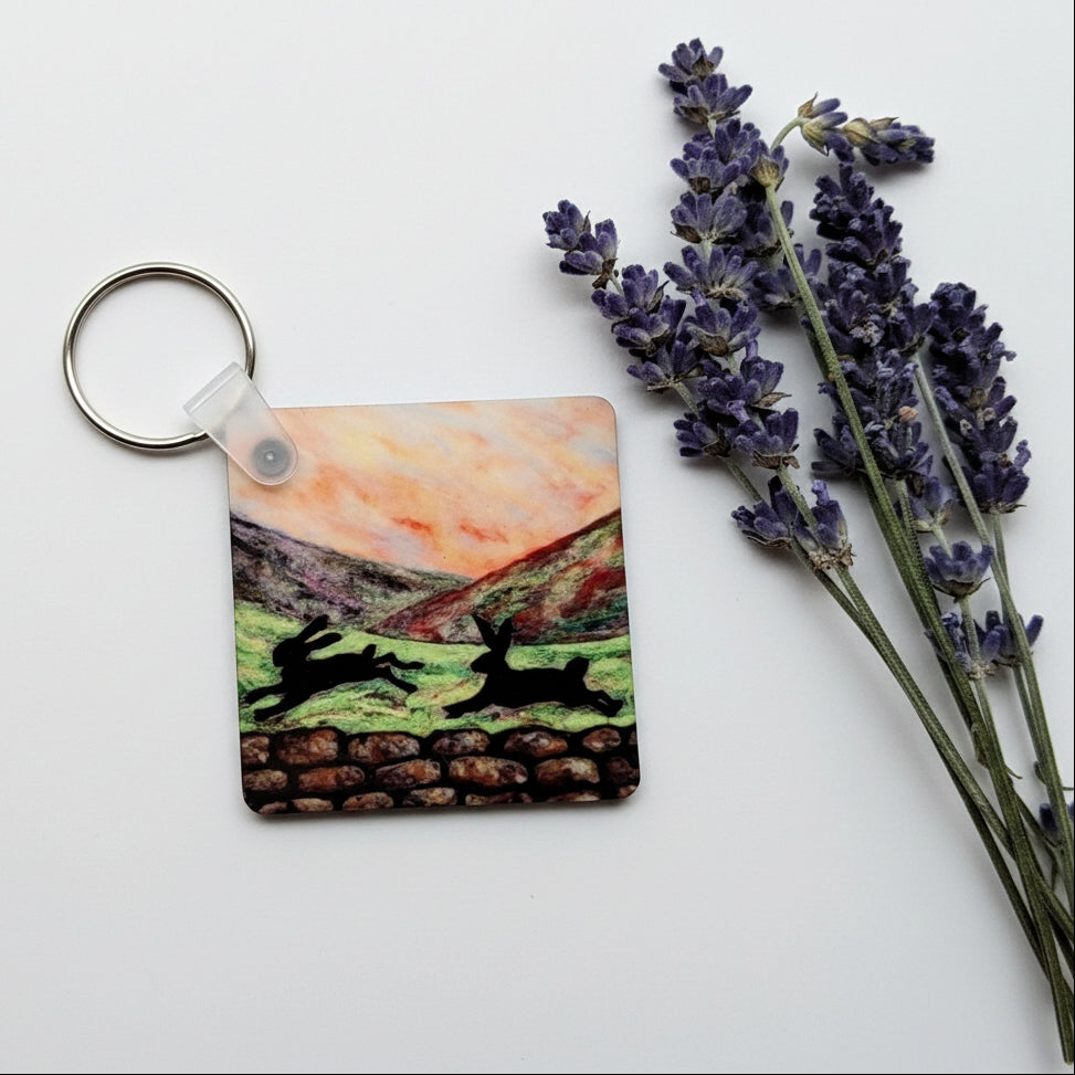 A square resin keychain featuring a painted landscape scene with deer, resting on a textured gray concrete surface next to dried lavender sprigs.