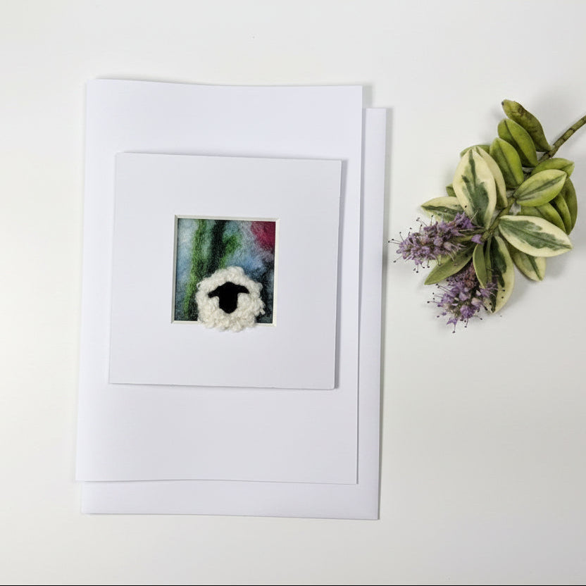 A square white art print or card featuring a small colorful painting of a sheep's head, displayed on a rustic whitewashed wooden surface next to a decorative green and purple plant sprig.