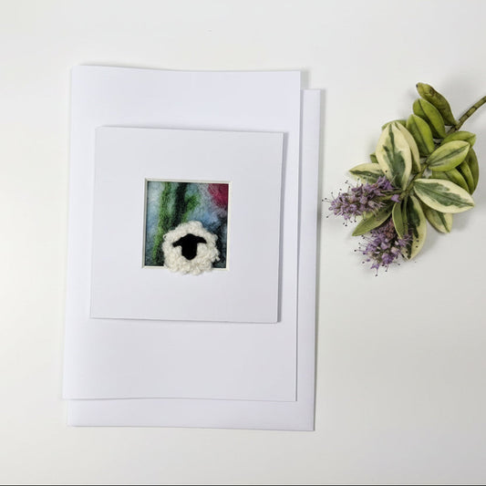 A square white art print or card featuring a small colorful painting of a sheep's head, displayed on a rustic whitewashed wooden surface next to a decorative green and purple plant sprig.