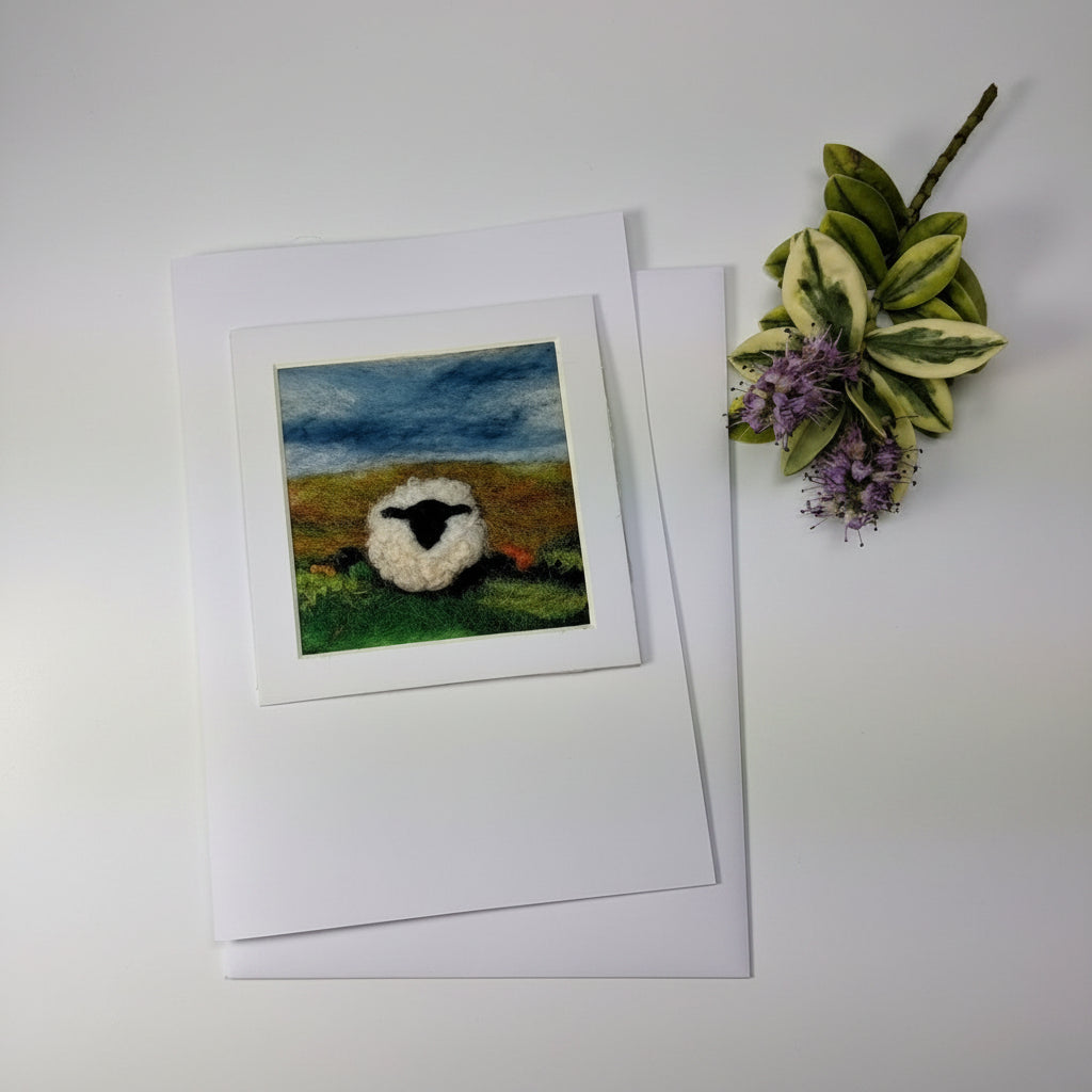 A square art print featuring a crocheted sheep in a field under a cloudy sky, displayed on a white card and envelope over a distressed white wood surface with a decorative leafy sprig.