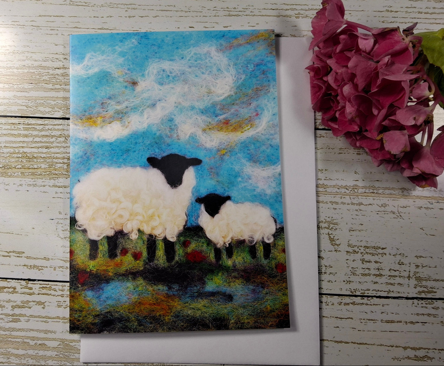 A square art print or card featuring a painted scene of two white sheep with black heads standing in a grassy field under a blue sky, placed on a white surface next to a pink flower.