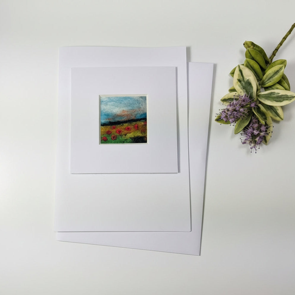 A matted art print or card featuring a small abstract painting with blue, green, red, and yellow tones, displayed on a rustic whitewashed wooden surface with dried flowers and a paintbrush nearby.