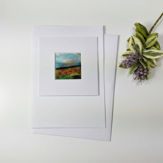 A matted art print or card featuring a small abstract painting with blue, green, red, and yellow tones, displayed on a rustic whitewashed wooden surface with dried flowers and a paintbrush nearby.