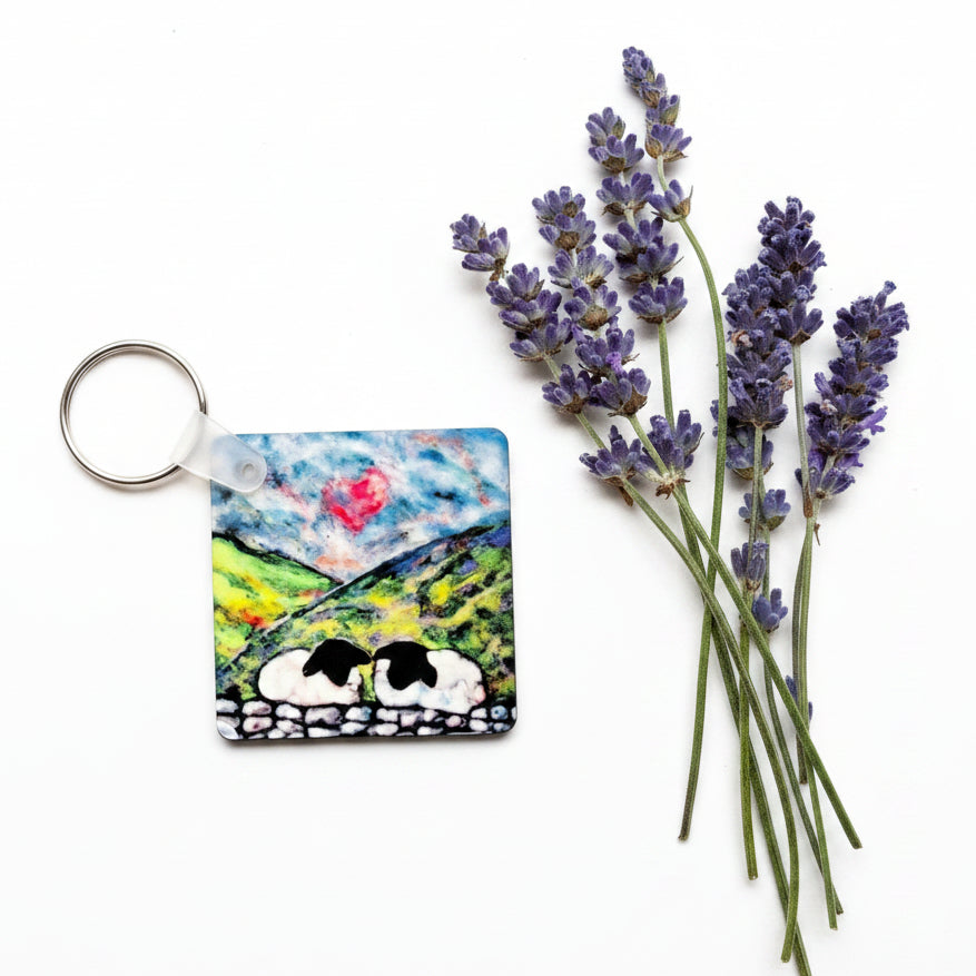 A square painted keychain with a metal split ring, featuring a colorful landscape scene with cows, placed on a textured gray surface next to lavender sprigs.