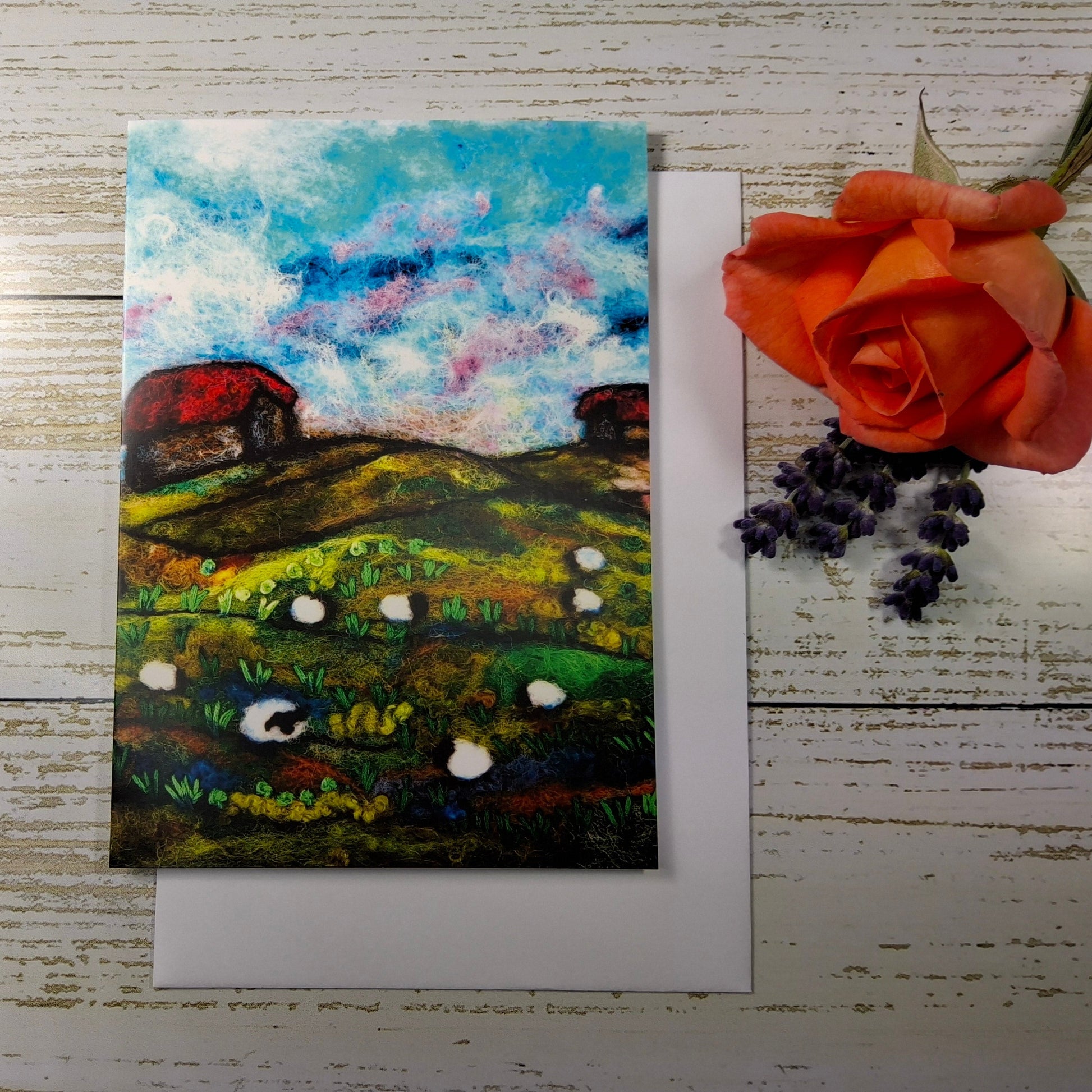 A square landscape art print or card featuring an impressionistic painted field with green hills, white sheep, red barns, and a blue sky,