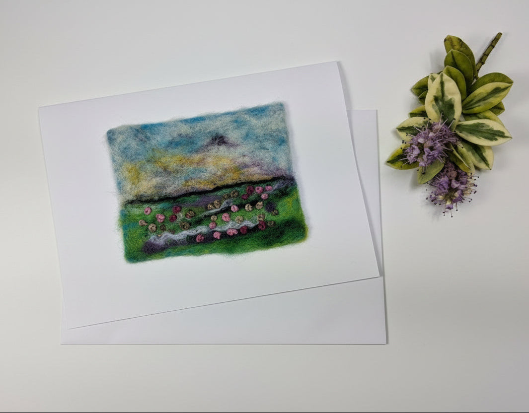 A small square landscape art print on white paper, featuring a painted green field with colorful flowers under a blue and yellow sky, displayed on a distressed white wood surface next to dried flowers.