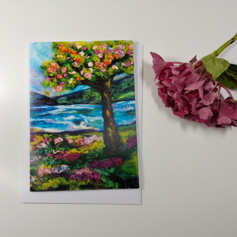 A square art print or card featuring a colorful painted tree with flowers and a blue ocean scene, placed on a white wood surface next to a pink hydrangea stem.