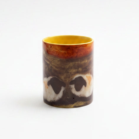 A ceramic mug with a yellow interior and handle, featuring a printed graphic of a raccoon face, sits on a gray stone countertop next to a small bunch of green herbs. A textured gray backdrop sheet is leaning against the wall behind the mug.