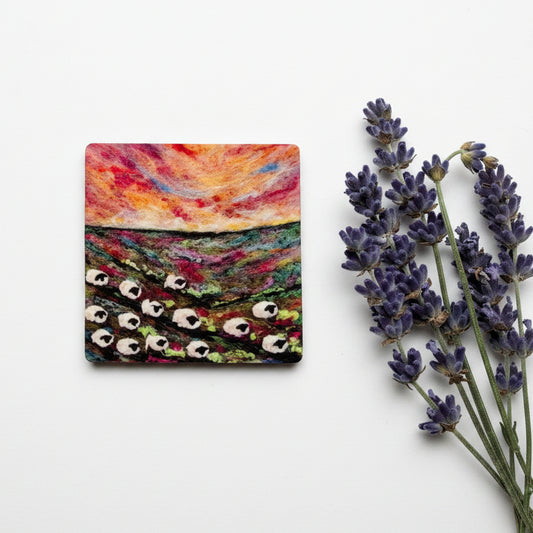 A small square painted card or coaster featuring an abstract landscape with a fiery orange and pink sky and a dark field with white flower-like shapes, placed on a gray textured surface next to lavender sprigs.
