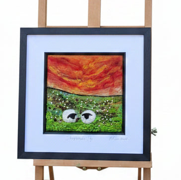 A framed art print featuring a split design of red/orange abstract shapes on the left and green grass with cartoon eyes on the right, displayed on a wooden easel outdoors next to a stone wall and trees.