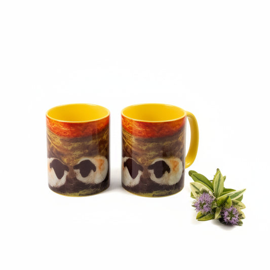 Two ceramic mugs with a brown and gold owl design and bright yellow interiors, displayed on a tabletop next to a lit studio light and a large marble-like column.