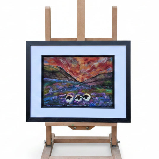 A framed landscape painting displayed on a wooden easel outdoors in a garden setting. The artwork depicts a dramatic sunset or sunrise over a lake with two small boats, surrounded by a white mat and black frame.