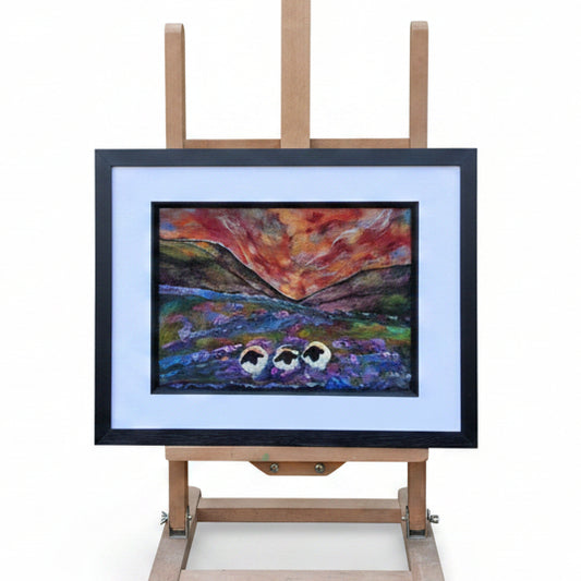 A framed landscape painting displayed on a wooden easel outdoors in a garden setting. The artwork depicts a dramatic sunset or sunrise over a lake with two small boats, surrounded by a white mat and black frame.