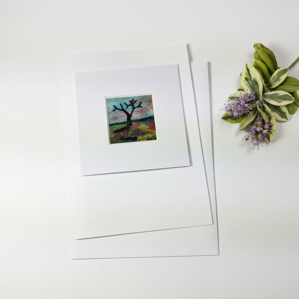 A matted art print featuring a small colorful square painting of a tree branch, displayed on a rustic white wood background with decorative greenery in the corner.