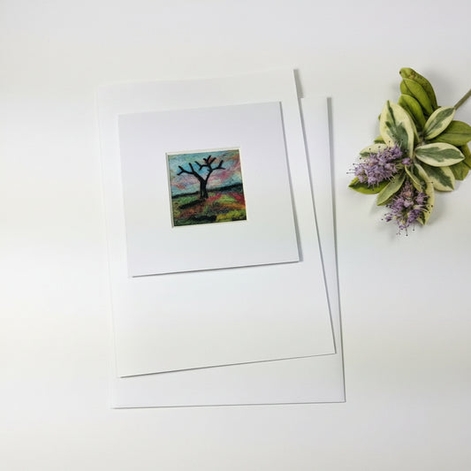 A matted art print featuring a small colorful square painting of a tree branch, displayed on a rustic white wood background with decorative greenery in the corner.