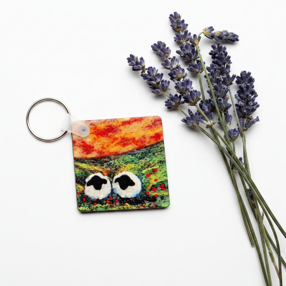 A small square keychain with a colorful landscape design and a silver key ring, styled on a textured gray concrete surface next to dried lavender sprigs.