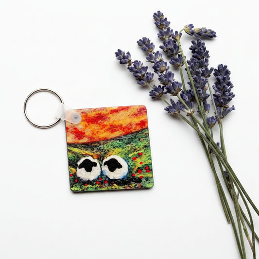 A small square keychain with a colorful landscape design and a silver key ring, styled on a textured gray concrete surface next to dried lavender sprigs.