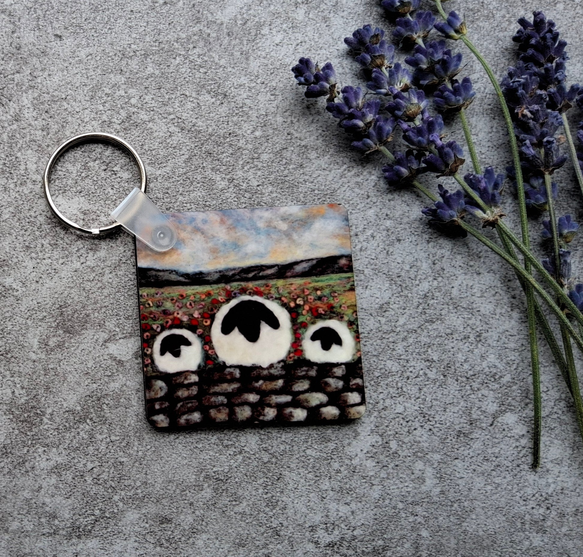 A square keychain with a silver split ring, featuring a printed design of three sheep on a hillside, styled on a textured gray surface next to sprigs of lavender.