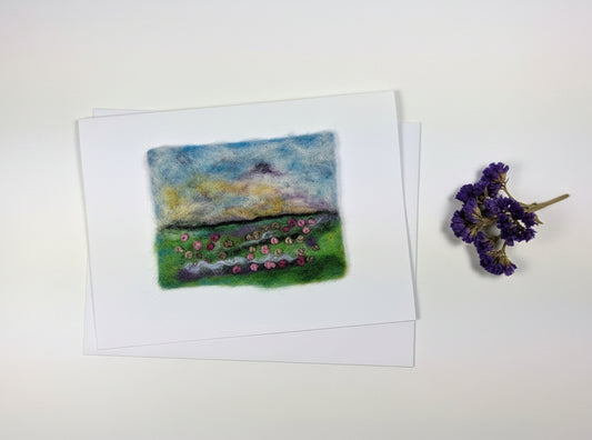 A handmade felt painting of a landscape with a blue and yellow sky, green hills, and small pink flowers, displayed on a white card over a rustic white wooden table.