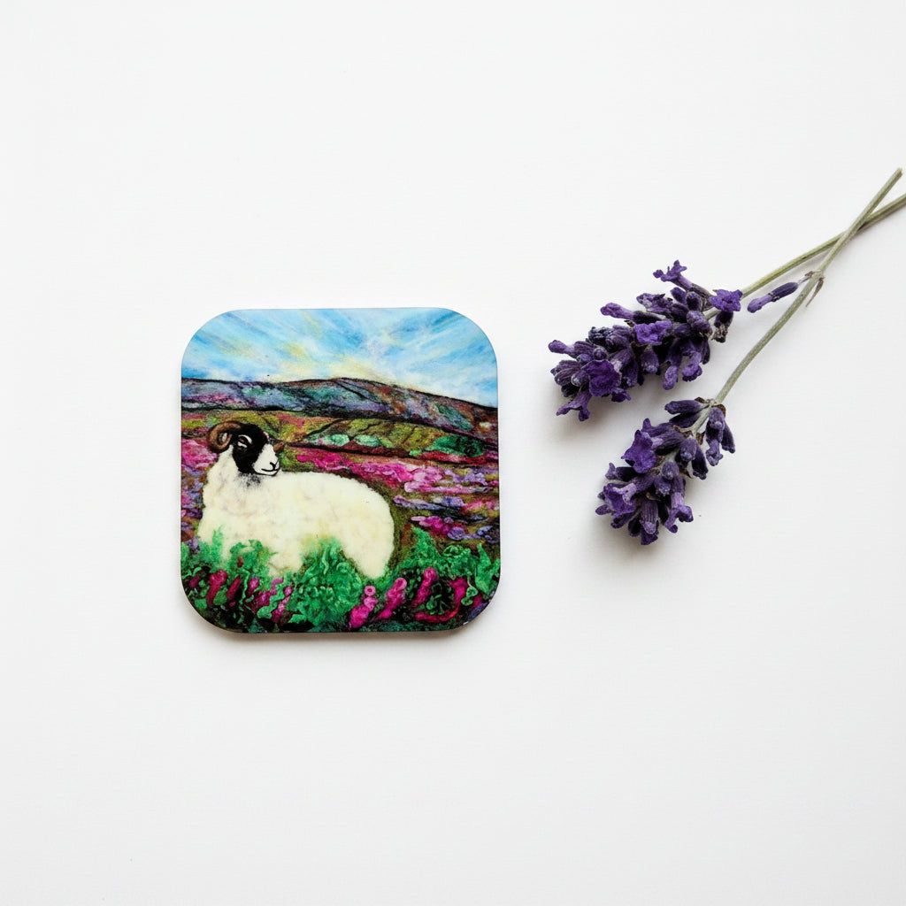 A square coaster with rounded corners featuring a colorful painted landscape of a white sheep in a field under a blue sky, displayed on a textured gray surface next to a sprig of purple lavender.