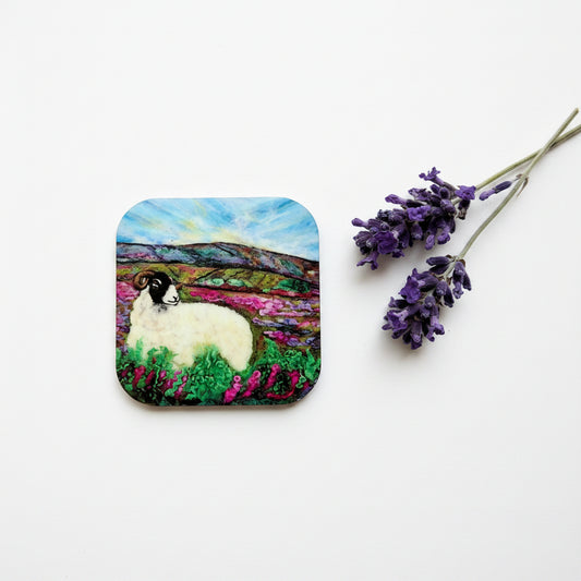 A square coaster with rounded corners featuring a colorful painted landscape of a white sheep in a field under a blue sky, displayed on a textured gray surface next to a sprig of purple lavender.