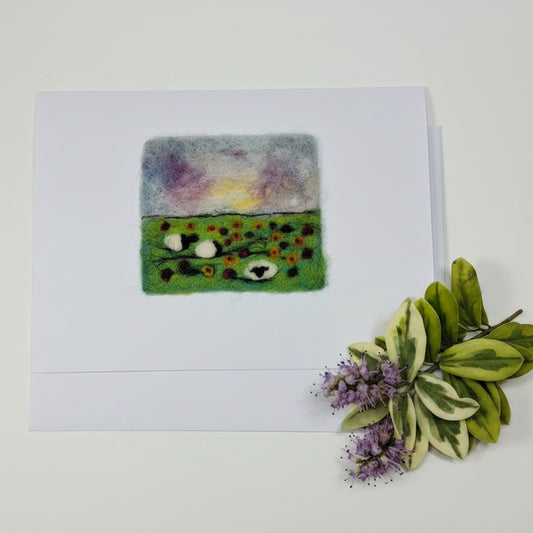A white square art print or card featuring a small felted wool artwork with a split design: one side is pastel purple, yellow, and blue, and the other is green with small colorful dots and white circular details. The print is photographed on a whitewashed wooden surface with dried flowers in the top corner.