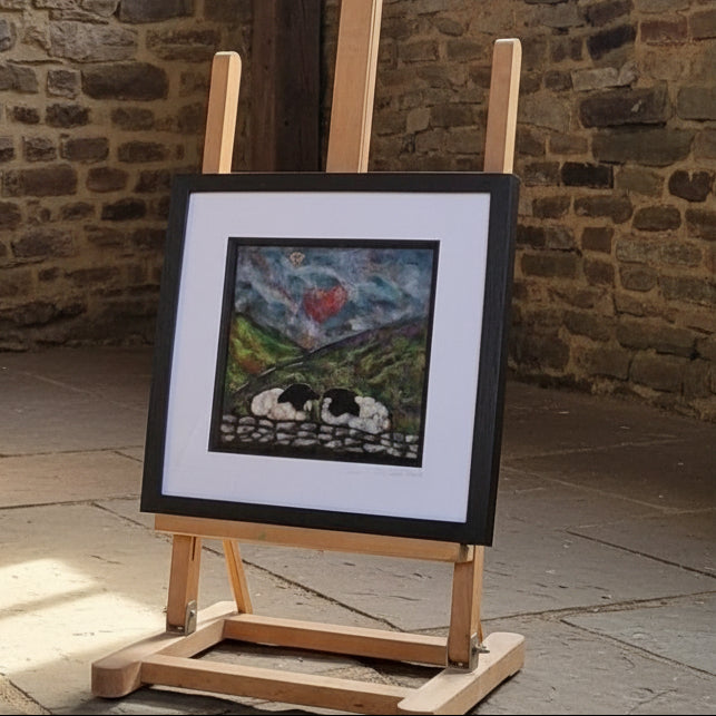 A framed art print or painting displayed on a wooden easel outdoors beside a stone wall and trees.