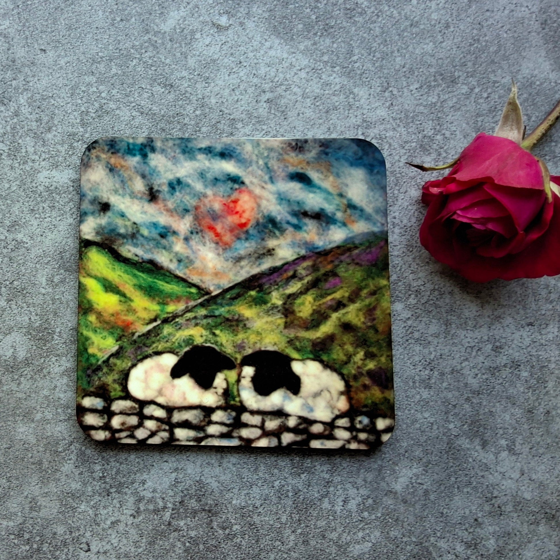 A square coaster featuring a hand-painted landscape scene of two sheep in a field under a cloudy sky, styled on a gray textured surface next to a single red rose.
