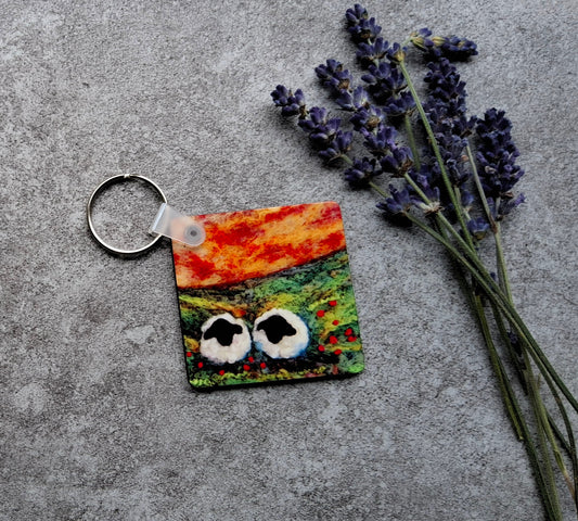 A square keychain with a colorful, abstract painted face design in orange, green, and red, attached to a silver metal key ring. It is styled on a textured gray concrete surface next to sprigs of dried lavender.