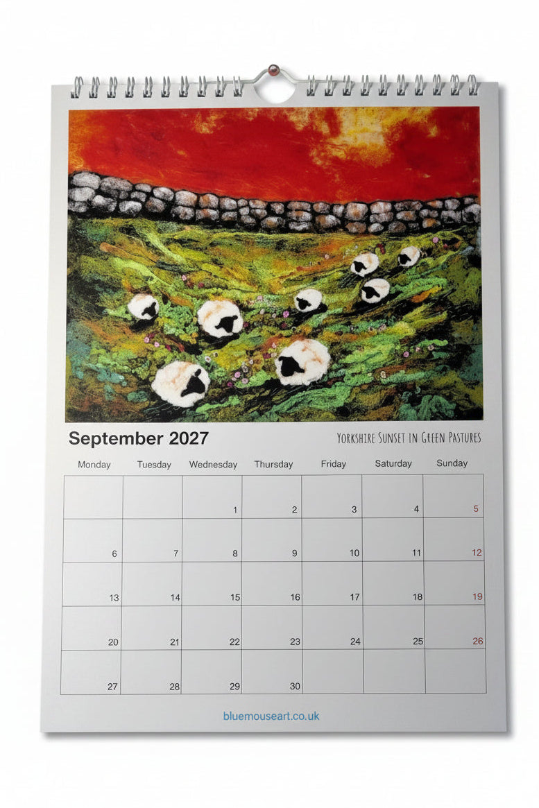 A spiral-bound September 2027 calendar page featuring a colorful painting of sheep on a green hill under a fiery red sky, photographed on a light wood surface.