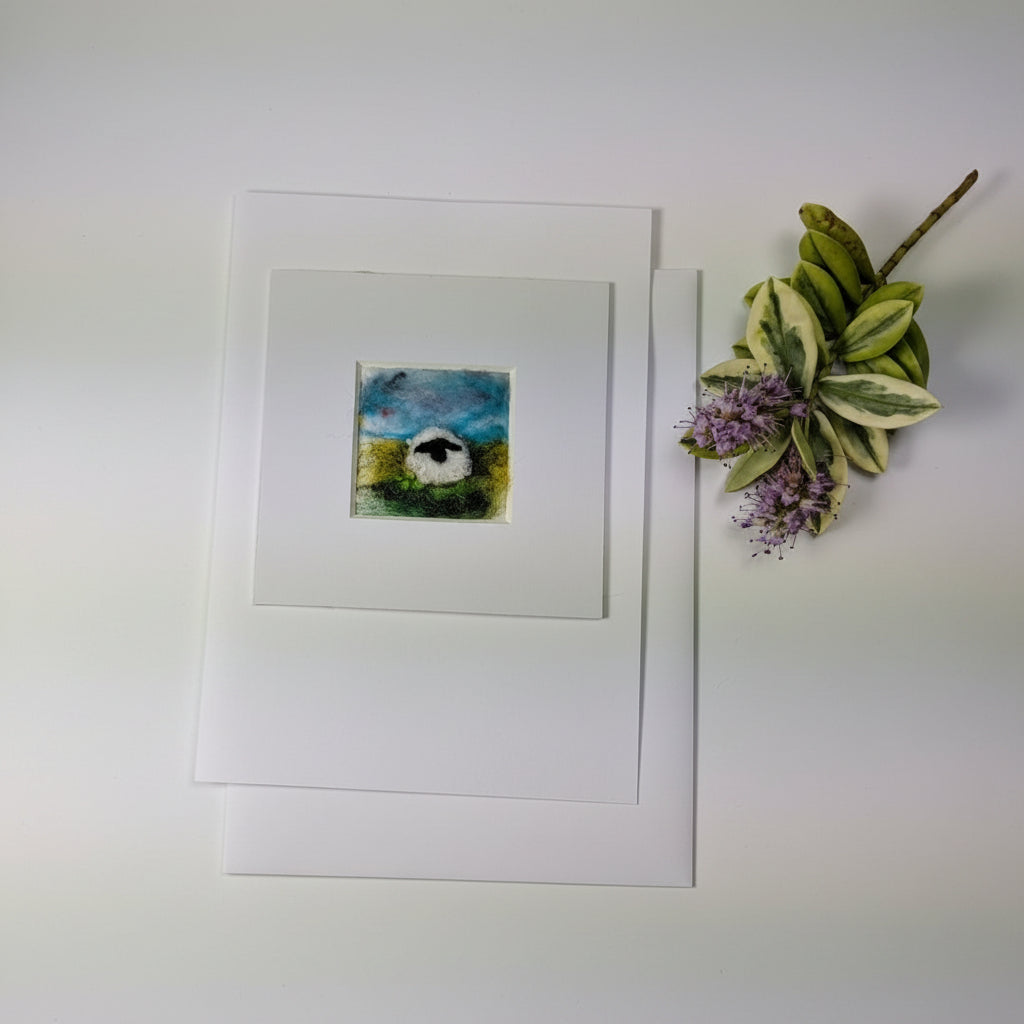 A square white art print or greeting card featuring a small colorful painting of a sheep in a field, displayed on a distressed white wood surface next to a decorative leafy sprig.