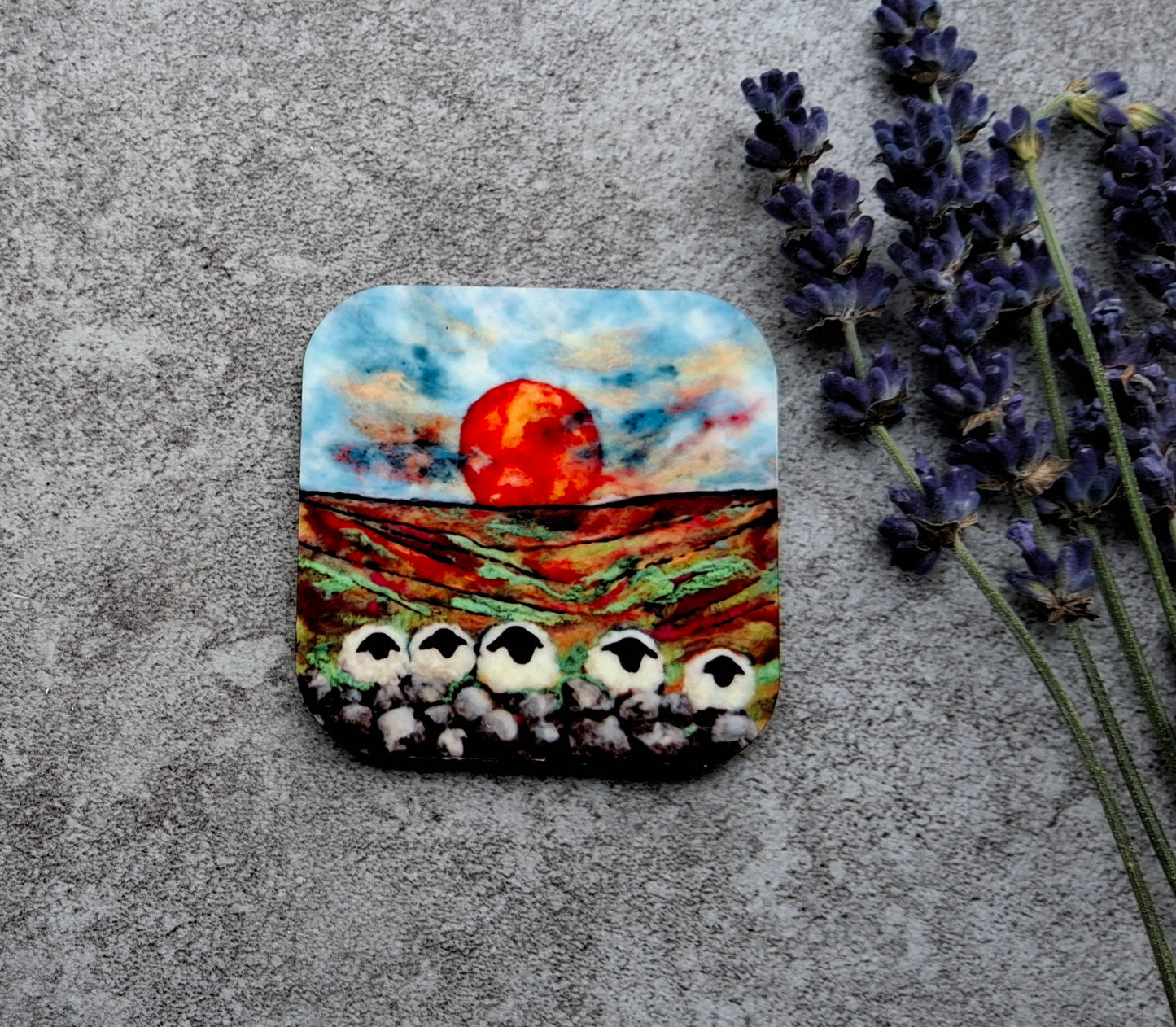 A square decorative plaque or coaster featuring a colorful landscape painting of a red sun over a field with white flowers, styled on a textured gray surface next to sprigs of lavender.