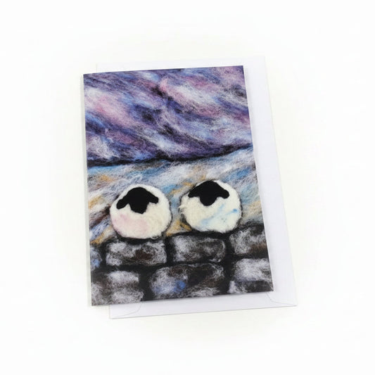 A square art print or card featuring a whimsical painting of two white penguins standing on a dark stone wall under a purple and blue sky, displayed on a white wood background next to a small sprig of purple flowers.