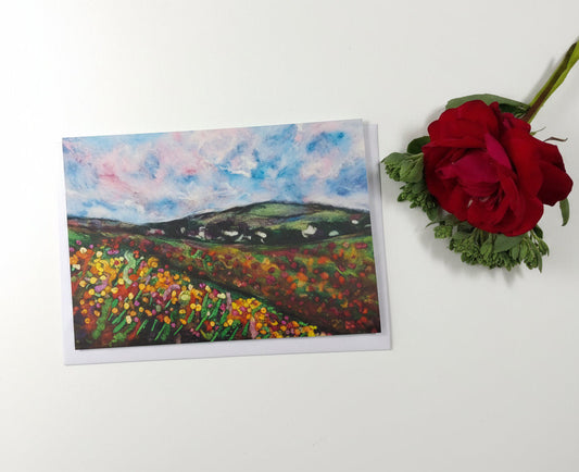 A landscape art print depicting a vibrant field of red and yellow flowers under a blue and pink sky, styled on a white wood background next to a red rose.