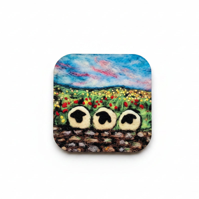A square painted stone or ceramic coaster with a landscape design of a blue sky, colorful flowers, and three owls, styled on a textured gray concrete surface next to sprigs of lavender.