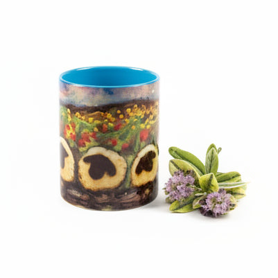 A cylindrical lamp with a light beige marble-look finish stands on a light-colored wooden desk next to a white adjustable desk lamp. In front of the lamp is a small patterned mug with a blue rim and a small sprig of greenery, with a dark room background.